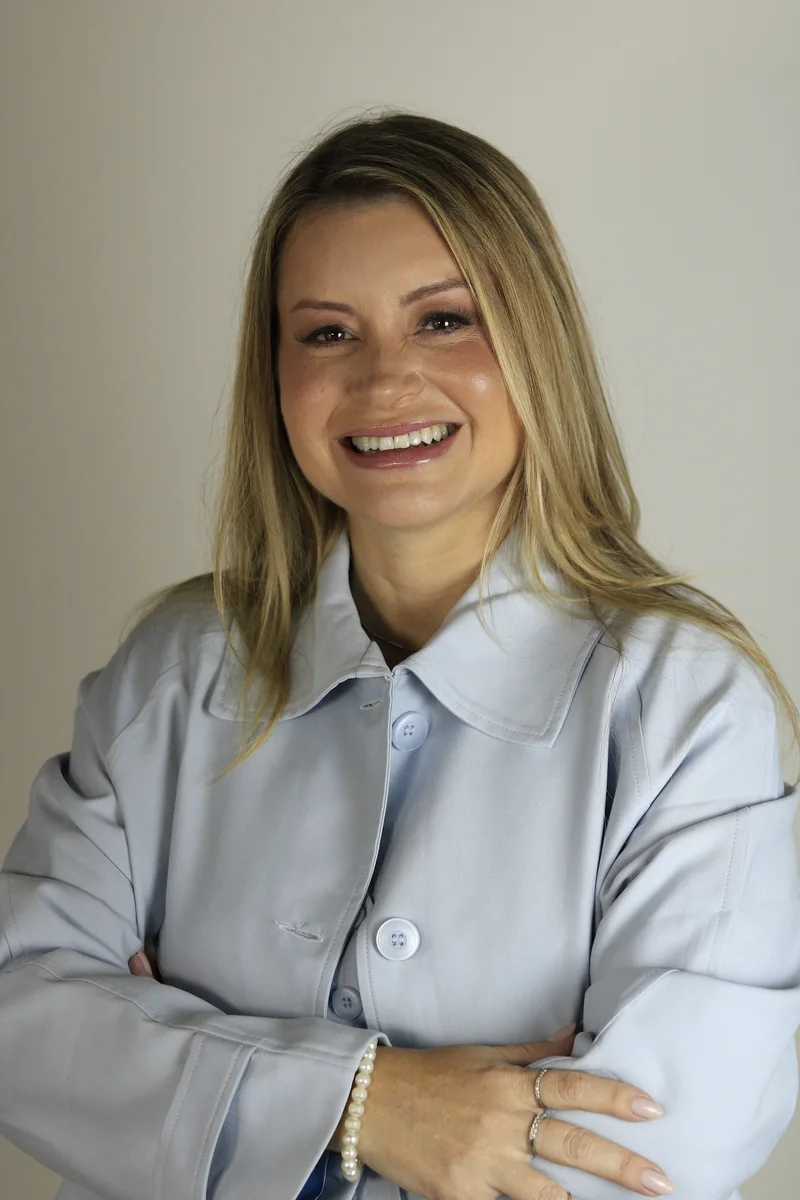Jaqueline Salles sorrindo, com blusa azul-clara e braços cruzados.